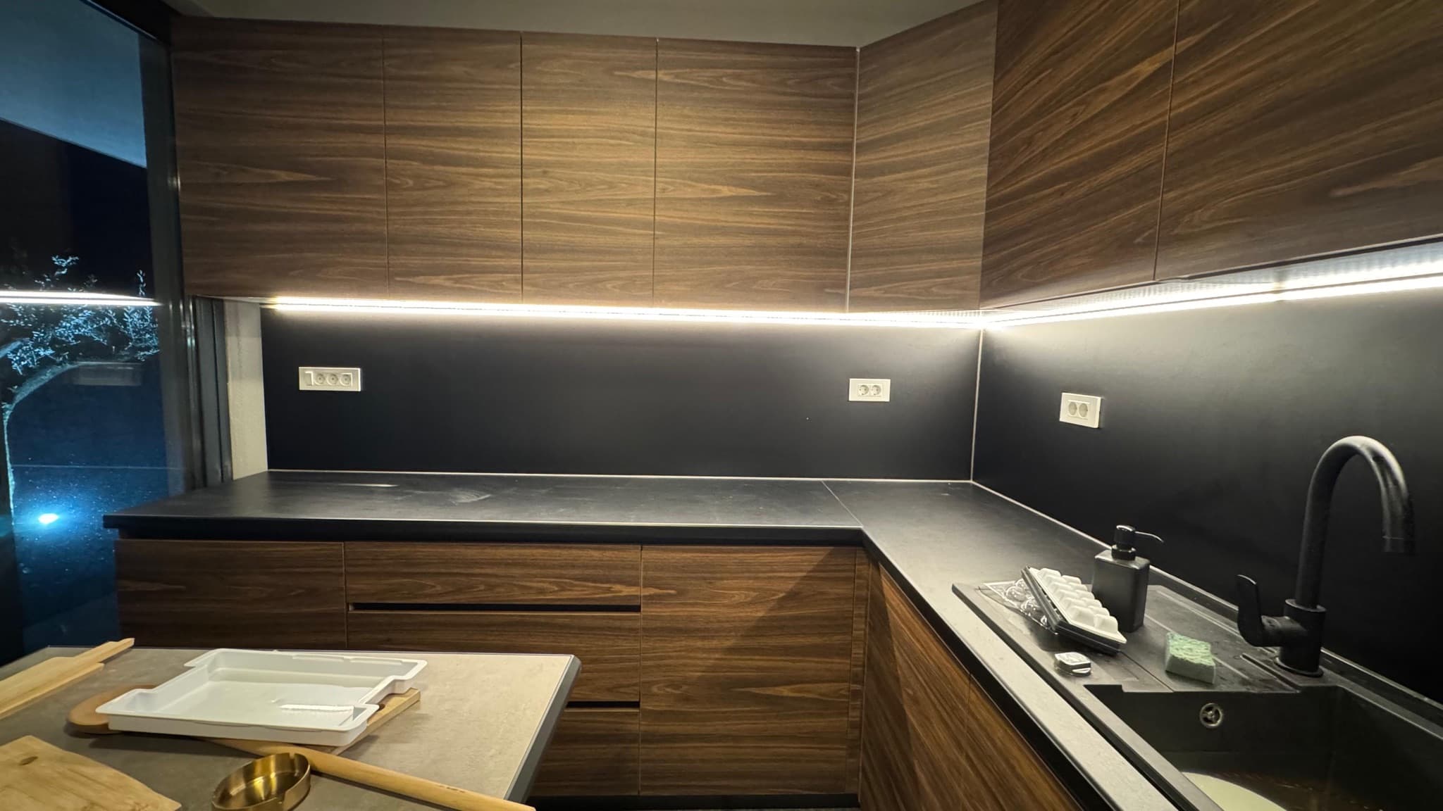 LED traka ispod visećih elemenata osvjetljava radnu plohu u kuhinji s tamnim furnirom.
